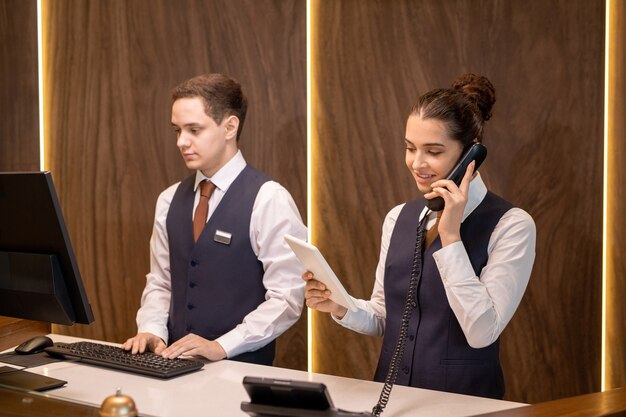 Photo one of two young hotel receptionists standing by counter, looking at touchpad display and consulting client on the phone against colleague