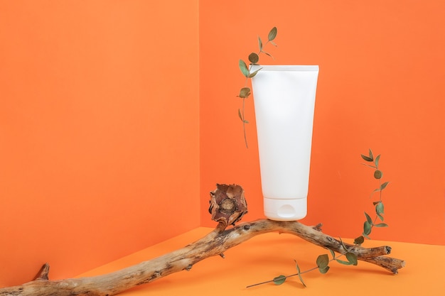 One white blank cosmetic tube bottle on wooden stick with dried flowers and eucalyptus branch in corner space on orange background. mockup front view copy space