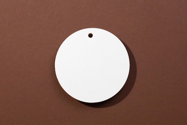 Одна белая картонная пустая бирка круглой формы с небольшим отверстием в верхней части, расположенная в самом центре на коричневом фоне.