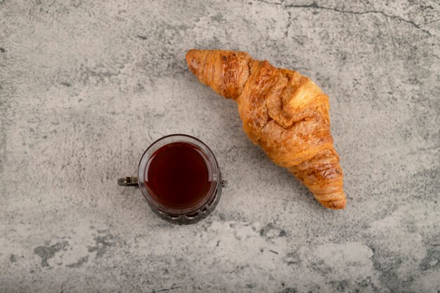 Photo one whole tasty buttery croissant with a glass cup of tea placed on a stone table .