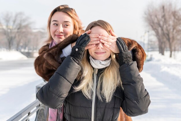 한 여성이 겨울 공원에 서 있는 다른 여성에게 손을 눈 뒤에 두었습니다