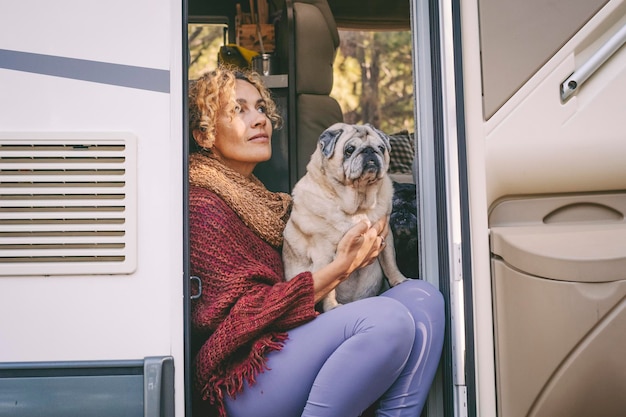 One woman sitting on the camper van door with old nice pug dog\
looking outside and enjoying relax and freedom female people living\
on a rv motorhome with animals and travel the world best\
friend