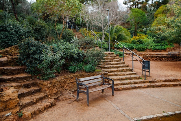 One wooden bench on alley among green garden at spring Guell park in Spain Walking path and stone steps leading to breathtaking viewpoint of Barcelona