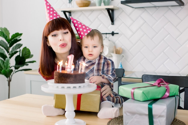 One year old baby girl blows out candles on cake on her birthday with her happy mom .