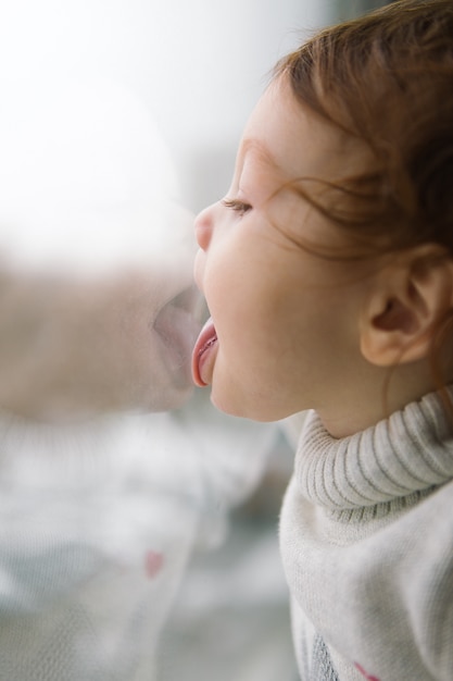 One year old baby. Little girl sitting on the window in a room. Children licking glass