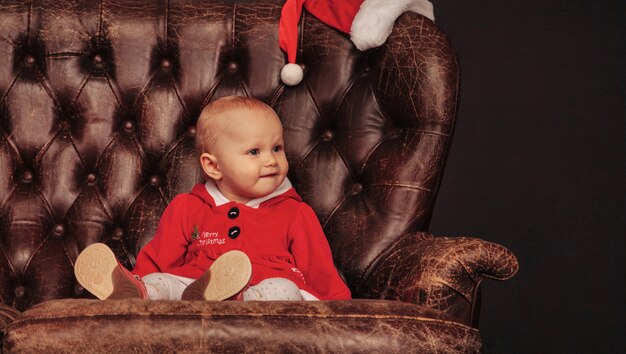 One year old blue eyes child in xmas costume on armchair in living room. Baby emotion on antique chair holiday evening. Concept of family celebration of Christmas and Happy New Year