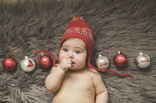 Una bambina di un anno giace su un copriletto, con un cappello rosso e gioca con i giocattoli di capodanno