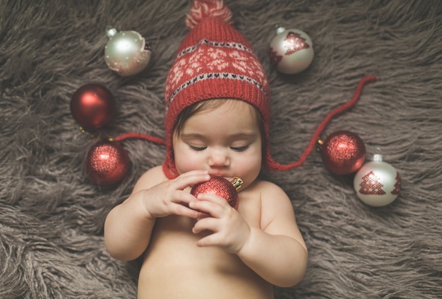 One-year-old girl lies on a bedspread, in a red hat and plays with New Year's toys