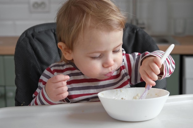 줄무늬 캐주얼 옷을 입은 한 살 배고픈 여자 아이는 하이체어에 있는 흰색 테이블에 앉아 숟가락으로 직접 죽을 먹습니다 흐릿한 식당 배경 아이들을 위한 건강한 식습관 아동 영양