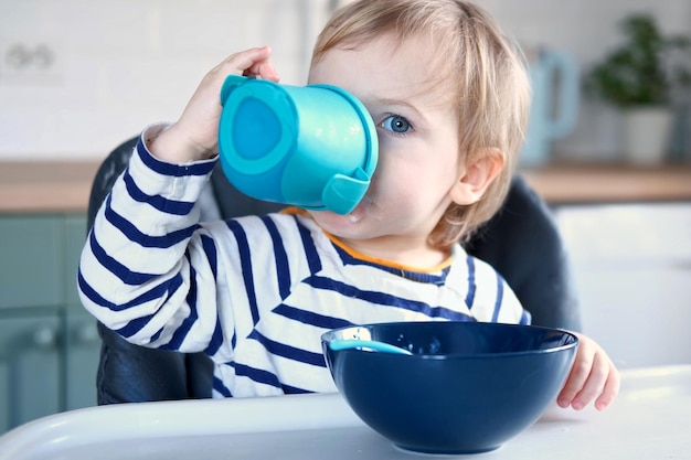 One year old hungry girl in striped casual clothes sits at white table in highchair drinks water from baby bottle sippy cup Blurred dining room background Healthy eating for kids Child nutrition