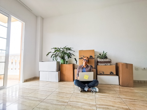 Photo one young beautiful woman in a empty house using her laptop with the boxes and packs