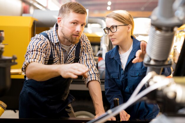 One of young confident technicians pointing at mechanism of industrial equipment during discussion of its qualities with colleague