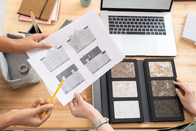 Photo one of young female designers or architects holding papers with house sketches while her colleague pointing at one of them