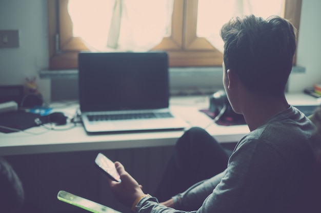 One young man using phone and looking at the laptop or computer at home in his room playing video games. Teenager addicted at social network and social media