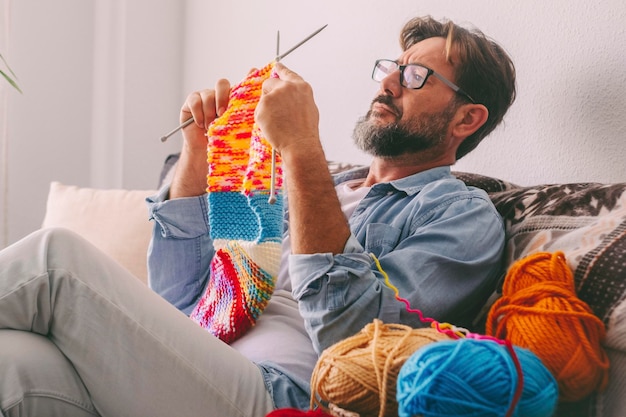 One young mature man knitting work with wool and needles at home sitting comfortable on the sofa in female indoor usual leisure activity Diversity and eguality female male People working indoor