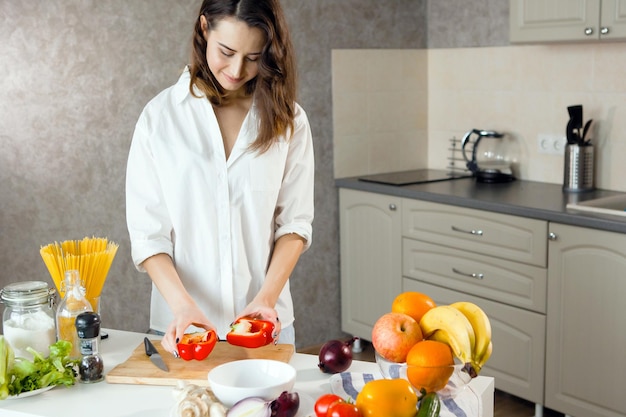 One young pretty woman in a white shirt in the kitchen cuts vegetables red sweet peppers corn blue onions Vegetarians healthy food