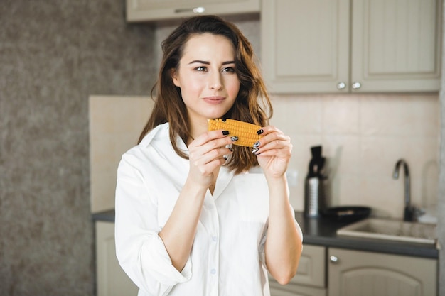 Photo one young pretty woman in a white shirt in the kitchen nibbles a corn cob