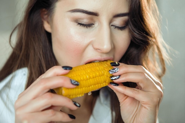 Foto una giovane donna graziosa con una camicia bianca in cucina mordicchia una pannocchia di mais