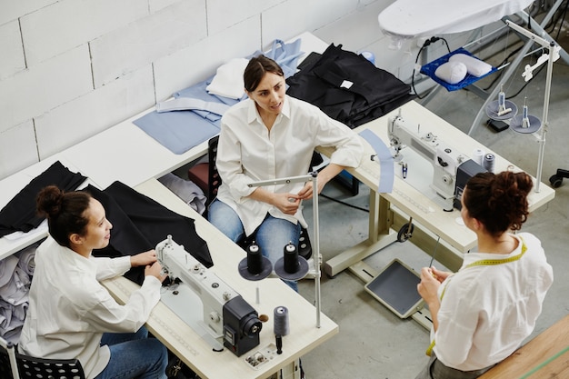 One of young seamstresses consulting her colleagues about details of new seasonal collection models and trends at meeting in workshop