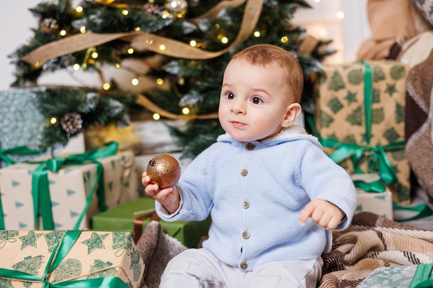 1 歳の男の子がプレゼントで飾られたクリスマス ツリーの近くに座っています 家の中のクリスマス ツリー 幸せな子供が新年を待っています