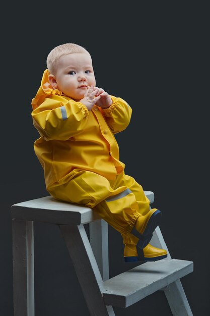 A oneyearold boy sits on the stairs in a yellow waterproof suit and looks at the camera