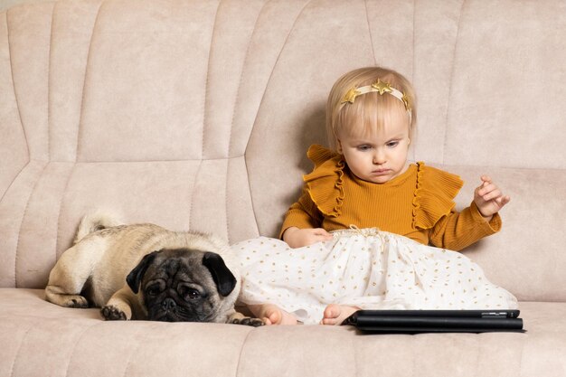 A oneyearold girl with a pug watches cartoons on a tablet sitting on the couch