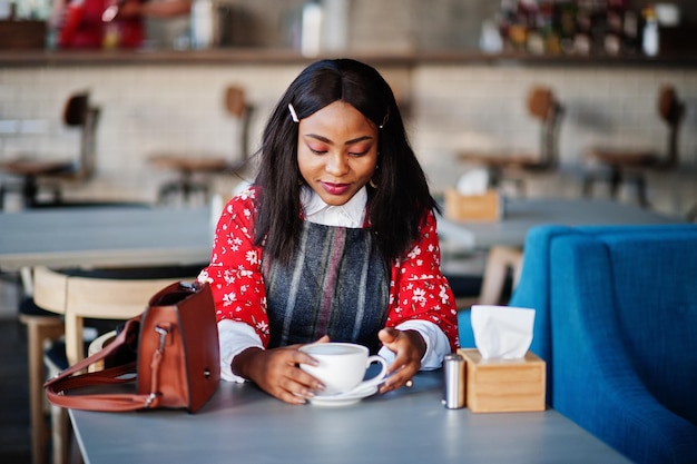 Сonfident young african american woman in smart casual wear at cafe with cup of hot drink in hand