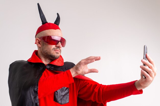 Foto ongebruikelijke blanke man in rode zonnebril, zwart rood halloween kostuum en hoed met duivelshoorns handgebaren tonen aan smartphone in zijn hand.