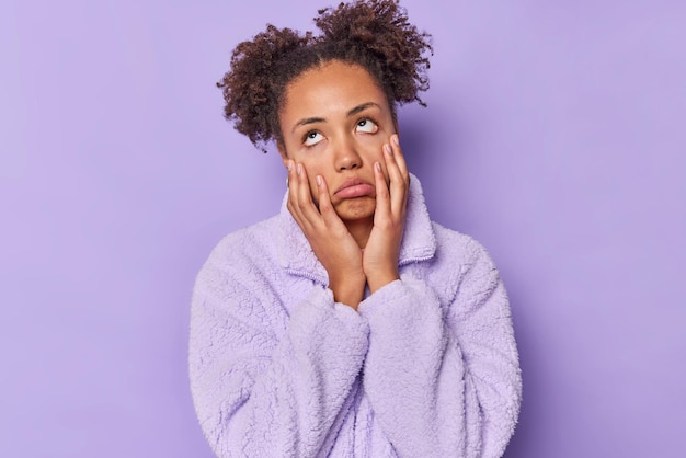 Foto ongeïnteresseerde verveelde jonge vrouw houdt handen op de wangen rolt ogen en voelt zich terughoudend toont apathie heeft krullend haar verzameld in twee broodjes gekleed in winterjas geïsoleerd over paarse achtergrond