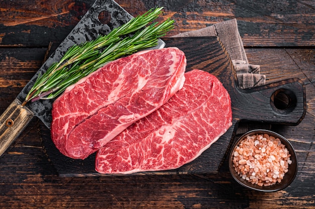 Ongekookt Raw Shoulder Top Blade of platte ijzeren rundvlees steaks op een houten slager bord met vlees hakmes. Donkere houten achtergrond. Bovenaanzicht.