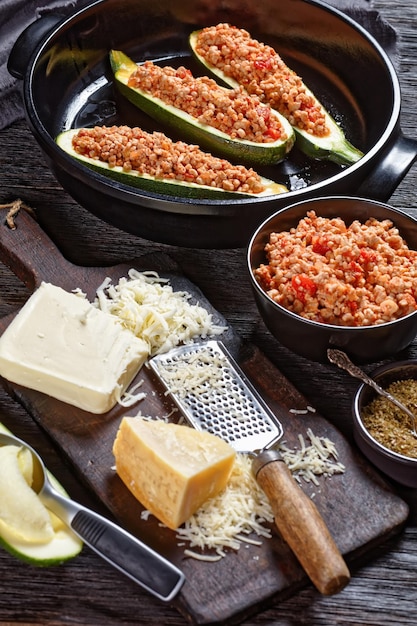 Ongekookte courgetteboten met gehakt, gehakte tomaten en kaas in een zwarte ovenschaal op een houten tafel met ingrediënten, Italiaanse keuken