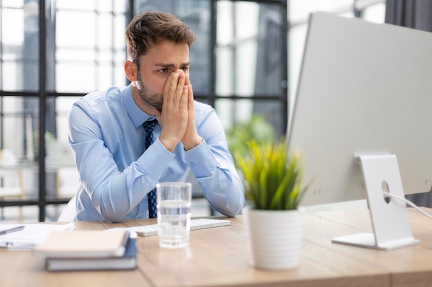 Ongelukkig gefrustreerd jong mannetje dat het hoofd vasthoudt door handen te zitten met de computer achter het bureau op kantoor