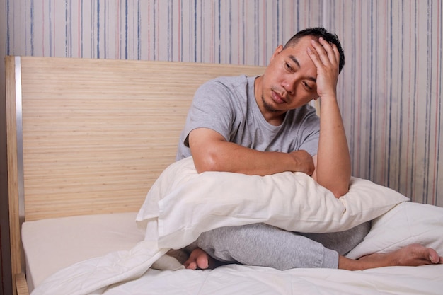 Ongelukkige Aziatische man die op bed zit en nadenkt over problemen die het gezicht met de handen sluiten