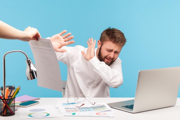 Ongelukkige, bebaarde man-analist verstopt zich voor extra werk op de werkplek met laptop, handen uitstrekkend om af te melden bij ontslag. Indoor studio-opname geïsoleerd op blauwe achtergrond