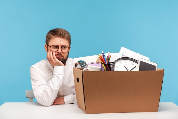 Ongelukkige depressieve man kantoormedewerker zittend op de werkplek met kartonnen doos met zijn dingen leunend hoofd op hand ontslag faillissement Indoor studio opname geïsoleerd op blauwe achtergrond