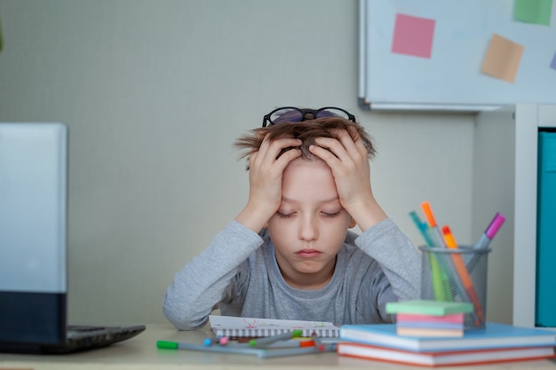 Ongelukkige en strssed schooljongen studeren met een notitieboekjes op zijn bureau thuis. Kind is moe van online leren. Onderwijs en kindertijd concept