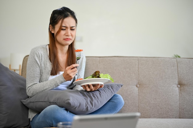 Ongelukkige en verdrietige jonge Aziatische vrouw is op dieet en probeert verse groenten of salade te eten