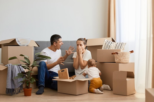 Ongelukkige gekke boze man die op zijn vrouw schreeuwt terwijl hij met haar in de buurt van de bank op de vloer zit en ruzie heeft in hun nieuwe appartement op de verhuisdag vrouw huilen