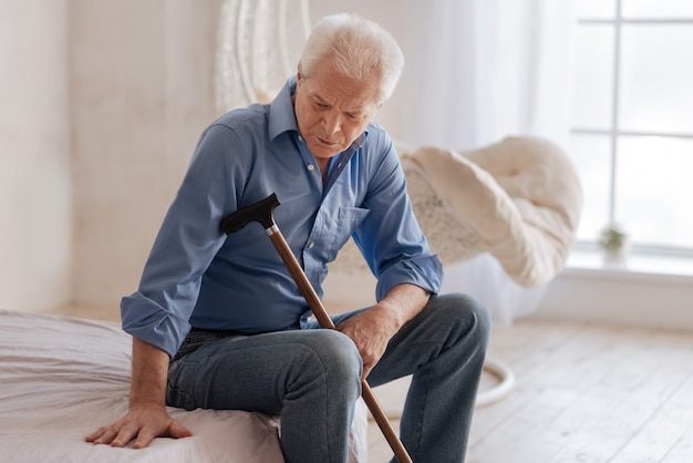 Ongelukkige humeurige bejaarde man die een wandelstok vasthoudt en naar de lege ruimte naast hem kijkt terwijl hij op het bed zit