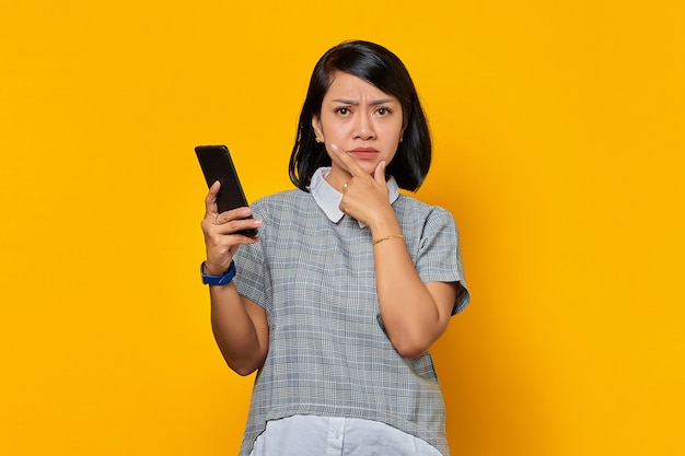 Ongelukkige jonge Aziatische vrouw met vinger op kin en met mobiele telefoon over gele achtergrond