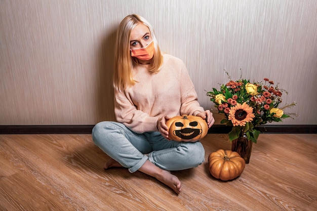 Ongelukkige jonge blonde vrouw met een medisch masker zit op de vloer en houdt een pompoen jack-o-lantern, herfstboeket met een zonnebloem in de buurt
