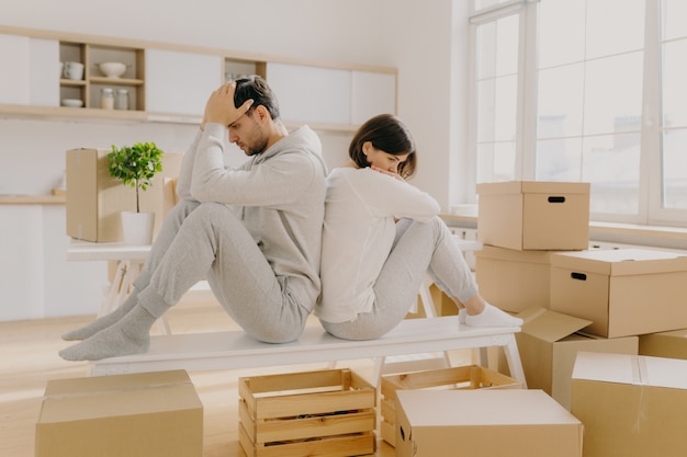 Ongelukkige jonge getrouwde vrouw en man moeten het huis verlaten, naar een andere plaats gaan, achterover leunen poseren in lege ruimte met stapel dozen, huiskleding en sokken dragen, hebben wat problemen