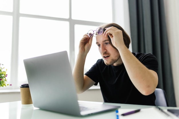 Ongelukkige jonge mannelijke werknemer met een bril kijkt naar het laptopscherm geschokt door defecte gadgets of operationele problemen gefrustreerde man verward verrast door onverwachte fout op computerapparaat