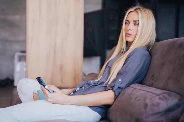 Ongelukkige jonge Scandinavische blonde vrouw in grijs shirt en witte broek zit op de bank en houdt telefoon vast