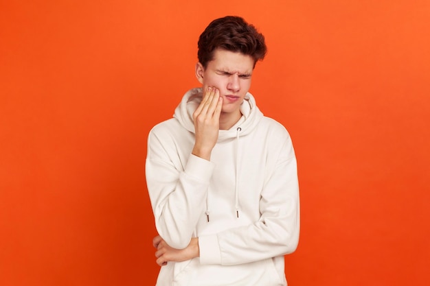 Ongelukkige jongeman in casual stijl sweatshirt met capuchon die lijdt aan vreselijke kiespijn tandvleesontsteking mondhygiëne stomatologie Indoor studio opname geïsoleerd op oranje achtergrond