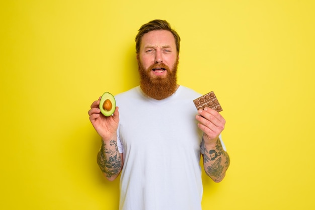 Ongelukkige man met baard en tatoeages houdt avocado en chocolade vast
