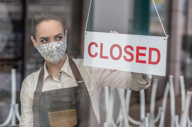 Ongelukkige serveerster die gezichtsmasker draagt, maakt een bord gesloten in een bar vanwege het coronavirus