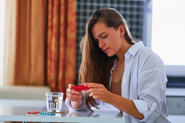 Ongezonde pijnlijke ongelukkige onwel vrouw die lijdt aan hoofdpijn en blisterverpakking met pijnstillerpillen thuis vasthoudt Gezondheidsproblemen