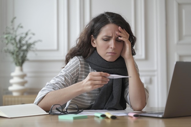 Ongezonde Spaanse vrouw gewikkeld in een warme gebreide sjaal die angstig op digitale thermometer kijkt