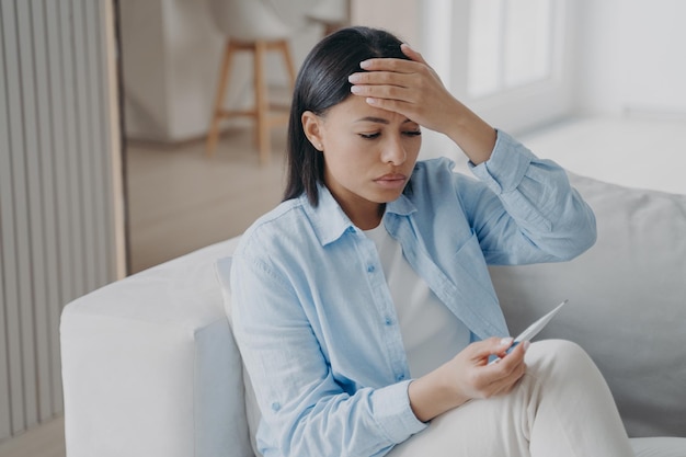 Ongezonde vrouw lijdt aan koorts controleer lichaamstemperatuur met thermometer zittend op de bank